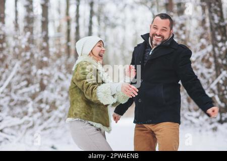 Bella coppia di famiglia felice di mezza età divertirsi all'aperto in vacanza invernale insieme, facendo una passeggiata attraverso la foresta innevata con sorriso, vestito Foto Stock