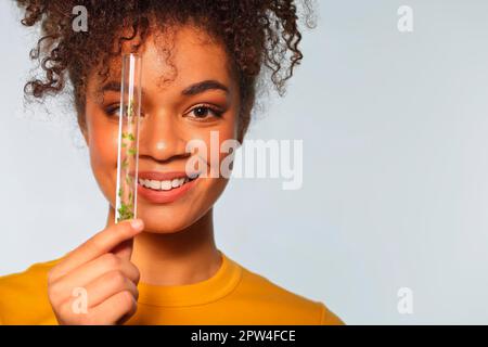 Donna afroamericana sorridente che tiene provetta di vetro con microgreens davanti al viso mentre posa isolato su sfondo grigio, copia spazio per Foto Stock