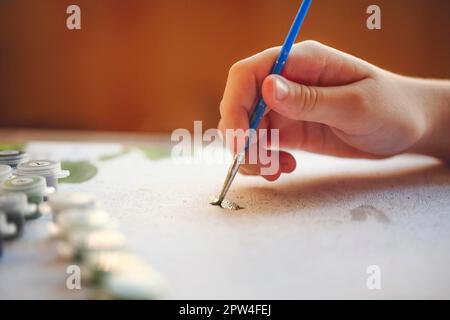 Foto ritagliata sfocata di pittura a mano femminile con pennello su carta, fuoco selettivo sulle vernici acriliche in scatole di plastica disposte in fila secondo Foto Stock