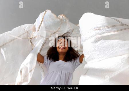 Sorridente ragazza afroamericana felice saltando con tessuto bianco in aria sullo sfondo grigio dello studio, sorridendo e ridendo ragazza nera dimostrare Foto Stock