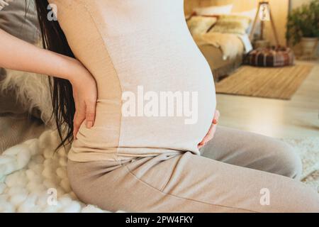 Primo piano foto di donna incinta che tiene il ventre mentre si siede sul divano a casa, donna che si aspetta il bambino toccare delicatamente il pancino, tagliato. Gravidanza Foto Stock