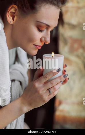 Ritratto laterale di giovane donna calma tenendo in mano la candela illuminata, chiudendo gli occhi avvicinandosi al viso e inalando il suo aroma, odorando familiare Foto Stock