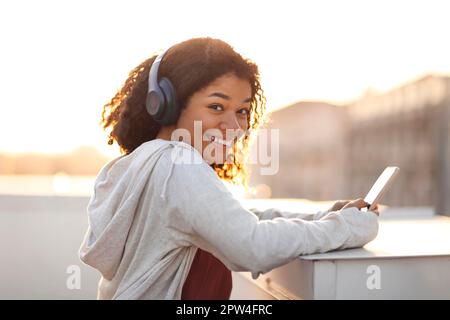 Vista laterale ritratto di felice ragazza afro-americana allegra in cuffie ascoltare musica su smartphone e sorridere alla fotocamera durante la mattina Foto Stock