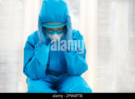 Medico stanco in dispositivi di protezione individuale PPE sensazione di depressione dopo un difficile spostamento stressante durante la pandemia di Covid-19, seduto a terra Foto Stock