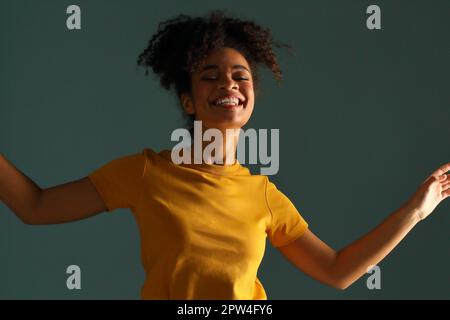 Bella donna di etnia afro-americana con pelle scura e bella in maglietta gialla che alza la mano nel saluto mentre guarda in macchina fotografica con un sorriso piacevole Foto Stock