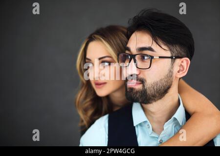 Giovane coppia etnica elegante che si abbraccia su sfondo grigio scuro parete guardando la fotocamera pensieroso. Ritratto della coppia Foto Stock