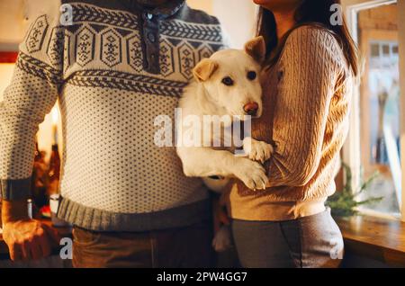 Giovane coppia felice uomo e donna che tiene carino cane bianco e abbraccio mentre in piedi in cucina decorata per Natale, bella famiglia con cucciolo Foto Stock