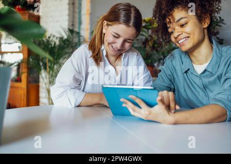 Due studenti di sesso femminile multietnica seduti al tavolo con un computer tablet, che parlano tra loro e si divertono. Istruzione, scuola superiore, persone e. Foto Stock