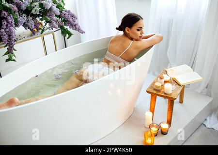 Vista dall'alto della giovane bruna che legge il libro e beve vino bianco in bagno con schiuma e candele Foto Stock