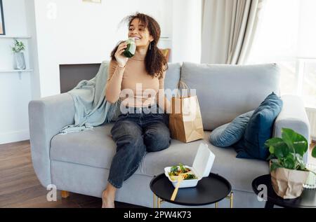 Donna afro-americana soddisfatta che mangia sana insalata di pasto equilibrata e verde fresco o frullato consegnato dal servizio di consegna del cibo. La donna a casa gode Foto Stock