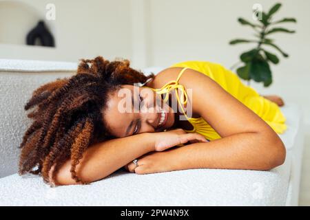 Bella curvy oversize giovane afto americano donna in un abito giallo si trova su un divano a casa. Corpo positivo elegante donna sorridente hanno tempo di relax. Foto Stock