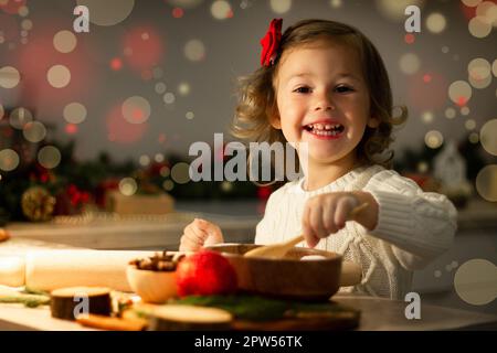 Carino piccola ragazza sorridente 2-4 con un arco rosso prepara i biscotti di pan di zenzero di Natale nella cucina del capodanno. Foto Stock