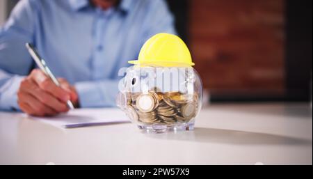 Cappello giallo duro su Piggy Bank trasparente su scrivania bianca Foto Stock