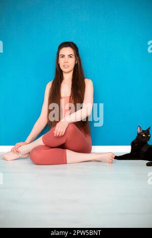 Giovane bruna donna che pratica yoga, seduta da parete blu a casa in mezzo loto esercizio con gatto nero a fianco, indossando bodysuit marrone e. Foto Stock