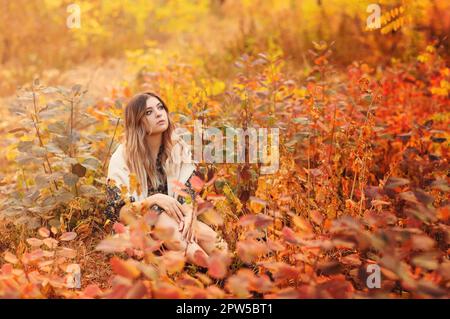 Bella giovane bionda donna che indossa stile boho vestito lungo mentre si siede con un look pensivo in erba alta nella foresta di autunno decorata in rosso e giallo Foto Stock