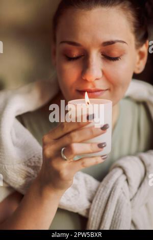 Ritratto laterale di giovane donna calma tenendo in mano la candela illuminata, chiudendo gli occhi avvicinandosi al viso e inalando il suo aroma, odorando familiare Foto Stock