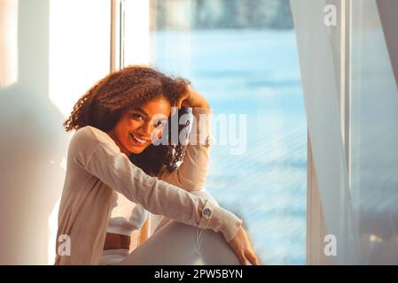 Felice African American ragazza etnia rilassato in abiti da casa seduti su davanzale sullo sfondo di tende che ondolano dal vento chiaro, felice afro Foto Stock