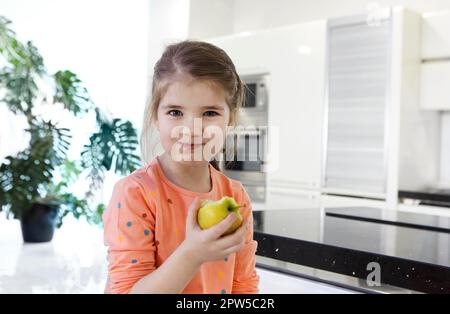 Piccola ragazza caucasica mangia una grande mela rossa in cucina. Bella bambina morde e mangia una mela. Guarda la fotocamera che tiene la frutta. Primo piano Foto Stock