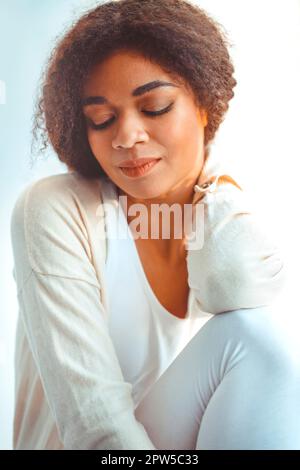Felice African American ragazza etnia rilassato in abiti da casa seduti su davanzale sullo sfondo di tende che ondolano dal vento chiaro, felice afro Foto Stock