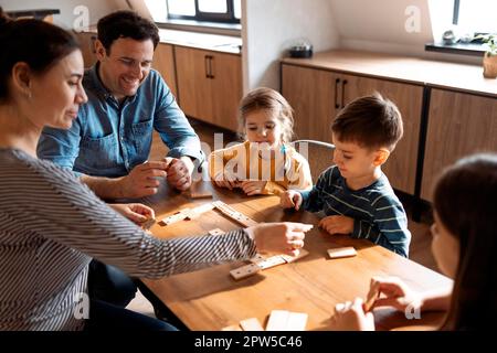 Madre, padre e bambini giocano a domino mentre si siedono nel soggiorno vicino alla finestra. Famiglia che passa il tempo insieme Foto Stock