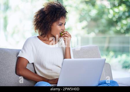 Giornata rilassata a casa con spuntini e tecnologia. una giovane donna che mangia una mela mentre usa il suo notebook a casa Foto Stock