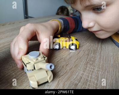 Serbatoio giocattolo su un tavolo di legno. Giocattolo di plastica. Artigianato da un designer per bambini. Il colore beige del serbatoio, immagine sfocata. Faccia e mano di un bambino. Foto Stock
