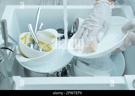 Foto ritagliata di una donna che indossa guanti monouso bianchi, lavando piatto bianco sporco con spugna sotto acqua corrente in lavandino bianco pieno di tovlewar sporco Foto Stock