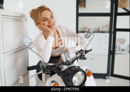 Ritratto di giovane carina donna motociclista in garage a casa. Donna millenaria sognante che guarda via sorridente pensando pensierosamente a fine settimana o vacanza Foto Stock
