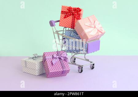 Piccole confezioni regalo di diversi colori con nastri risiede nel carrello spesa su un viola e blu sullo sfondo color pastello. Foto Stock