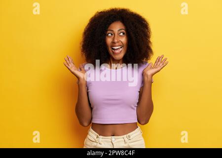 Donna spensierata positiva con capelli soffici posa a bocca aperta con sorriso gioioso, spalma palme e guarda da parte con espressione eccitata, si alza su y Foto Stock