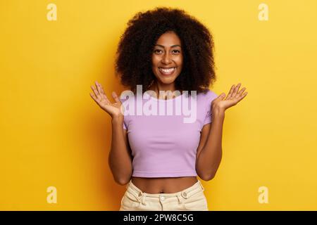 La giovane donna soddisfatta sorride felicemente, distende le palme da parte e guarda alla macchina fotografica, vestita con T-shirt viola e jeans, che si posa su sfondo giallo Foto Stock