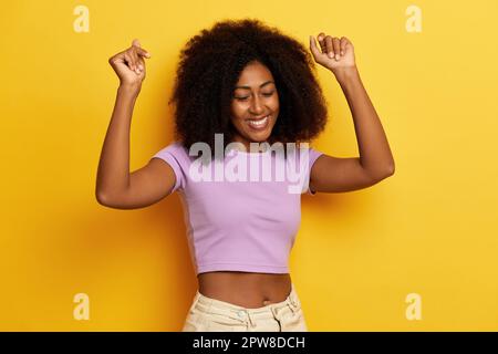 Foto di felice danza scuro pelle attraente donna con le mani sollevate, celebra il suo successo con sorriso soddisfatto, vestito in casual t-shirt viola Foto Stock