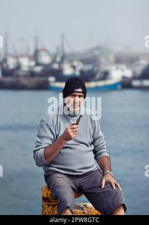 Pescatore, uomo maturo ritratto e fumo tubo vicino al mare in un porto per la pesca. Anziano, oceanico e lavoratore senior seduto in barca all'aperto Foto Stock