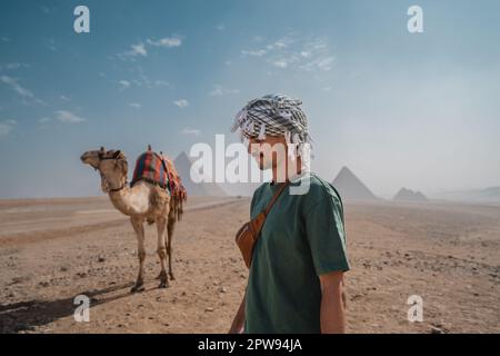 giovane in una turbante accanto ad un cammello con le piramidi sullo sfondo. El Cairo. Egitto Foto Stock