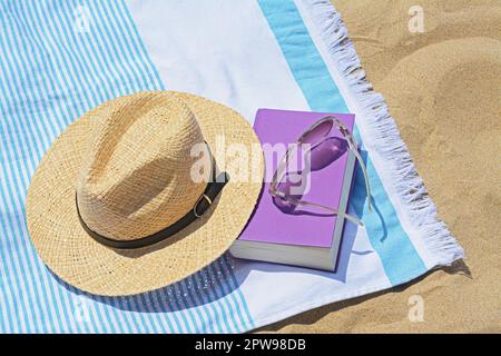 Telo da spiaggia con libro, cappello di paglia e occhiali da sole sulla sabbia Foto Stock