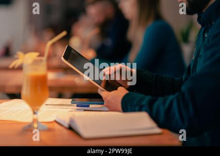 Primo piano di un uomo che tiene in mano un tablet, bevande e libri sul tavolo al bar Foto Stock