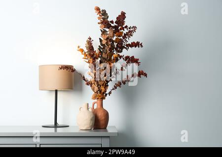 Vasi eleganti, rami di eucalipto secco e lampada da tavolo sul petto vicino parete bianca al chiuso, spazio per il testo. Design degli interni Foto Stock