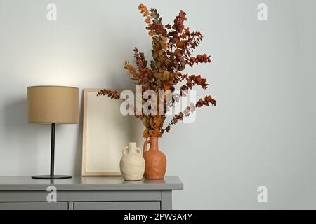 Vasi eleganti, rami di eucalipto secco e lampada da tavolo su cassettiera vicino parete bianca interni, spazio per il testo. Design degli interni Foto Stock