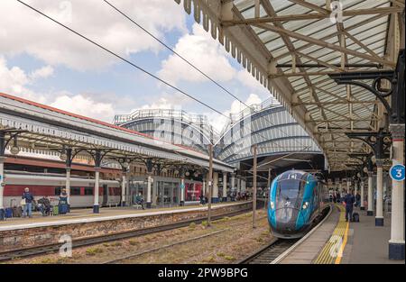 Un treno è parcheggiato lungo una piattaforma di una stazione con architettura del 19th ° secolo. Il passeggero aspetta i treni e un cielo con le nuvole è sopra. Foto Stock