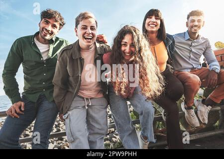 Giovani amici diversi abbracciando e ridendo, tenendo su una ringhiera sul mare, godendosi il loro tempo insieme. Foto Stock