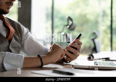Vista in primo piano delle mani maschili che toccano il telefono cellulare. Uomo di mezza età che utilizza lo smartphone mentre è seduto alla sua scrivania in un ufficio moderno. Attenzione selettiva a portata di mano Foto Stock