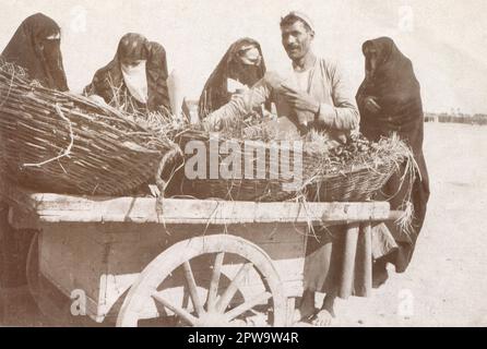 Egitto. Circa 1918. Un gruppo di donne sono raccolte intorno ad un venditore di strada che indossa una gallibaya, che sta vendendo le date dai cestini messi su un carrello gommato. Sta servendo il frutto in coni di carta arrotolata. Le donne indossano al-habara, composta da una gonna a tutta lunghezza, una copertura per la testa, e al-burqu’, una copertura per il viso. Due della donna indossano anche Arūsat al Burqa, ornamenti indossati sul velo facciale. Foto Stock