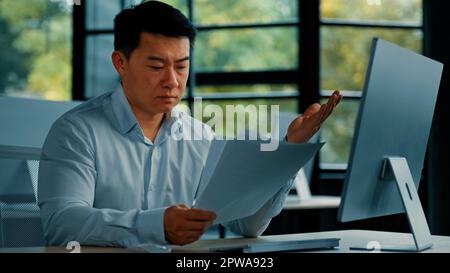 Disperato triste asiatico giapponese senior specialista businessman siede in ufficio leggere documenti truffa errore di notifica nella documentazione commerciale uomo ricerca Foto Stock