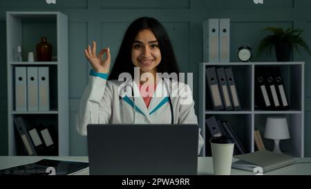 Felice positivo giovane donna medico arabo indiano etnia terapista medico guardando fotocamera sorridente fare bene mano gesto raccomandare bene Foto Stock