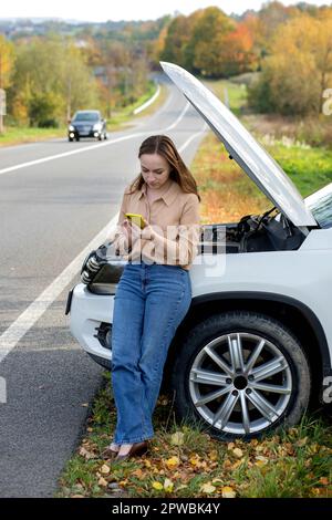 Una donna attende assistenza vicino alla sua auto rotta sul lato della strada. Donna con un'auto rotta con cofano aperto. Turbato giovane donna con cellulare vicino Foto Stock