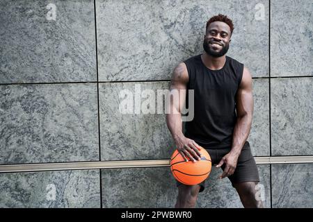 Forte giovane uomo nero sportivo che tiene la palla da basket in piedi all'aperto. Foto Stock