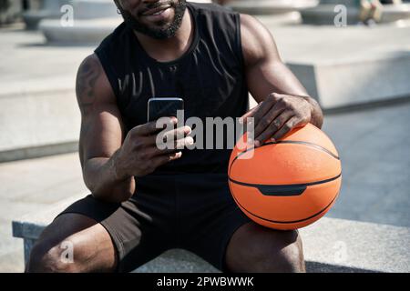 Indossate un giovane uomo nero sportivo che tiene la palla da basket utilizzando il telefono all'aperto. Foto Stock