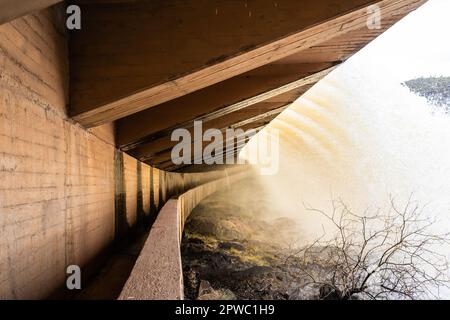 Una diga con le sue porte di alluvione aperte e un grande volume di acqua che scorre sulle rocce con le nuvole di tempesta. Foto Stock