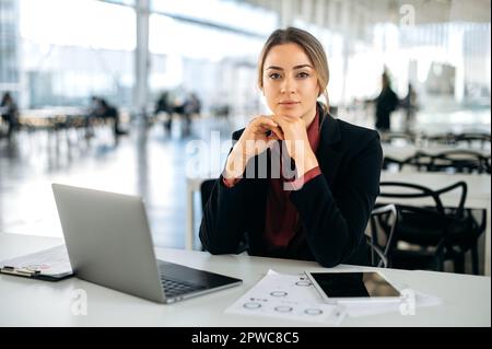 Bella fiduciosa influente donna d'affari caucasica, in elegante vestito, direttore della società, si siede su una scrivania in ufficio, guarda la fotocamera con un lieve sorriso Foto Stock