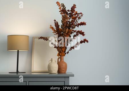 Vasi eleganti, rami di eucalipto secco e lampada da tavolo su cassettiera vicino parete bianca interni, spazio per il testo. Design degli interni Foto Stock